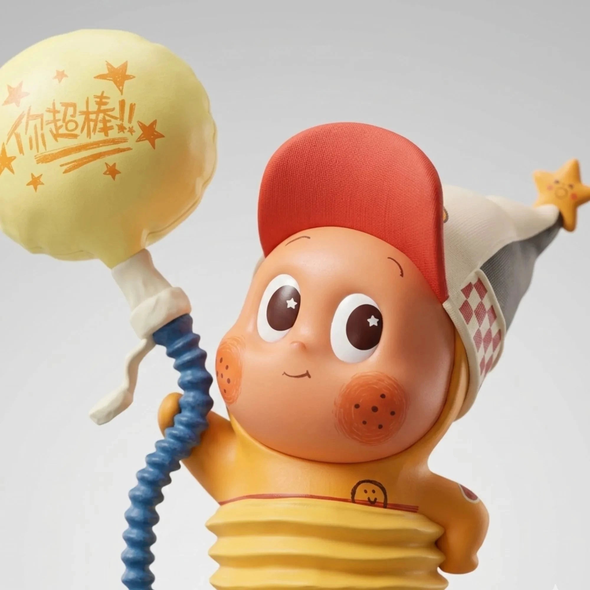 Un gros plan détaillé sur le visage de la figurine 'Little Balloon' de la collection Star Kid. Elle porte une casquette de baseball rouge et un chapeau de magicien gris à carreaux. La main gauche de la figurine est visible, tenant un tuyau bleu enroulé et un grand ballon jaune en forme de bulle de texte avec l'inscription '你超棒!!' et des étoiles en orange. Le fond est un studio gris clair et uni.