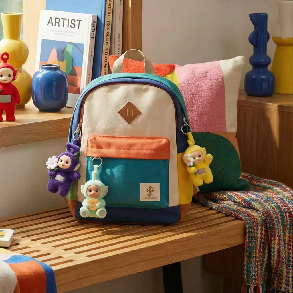 Sac à dos en toile blanc avec poches bleu et jaune, orné de charms Teletubbies (Tinky Winky, Dipsy, Laa-Laa, Po), posé sur un banc en bois dans un intérieur stylisé.